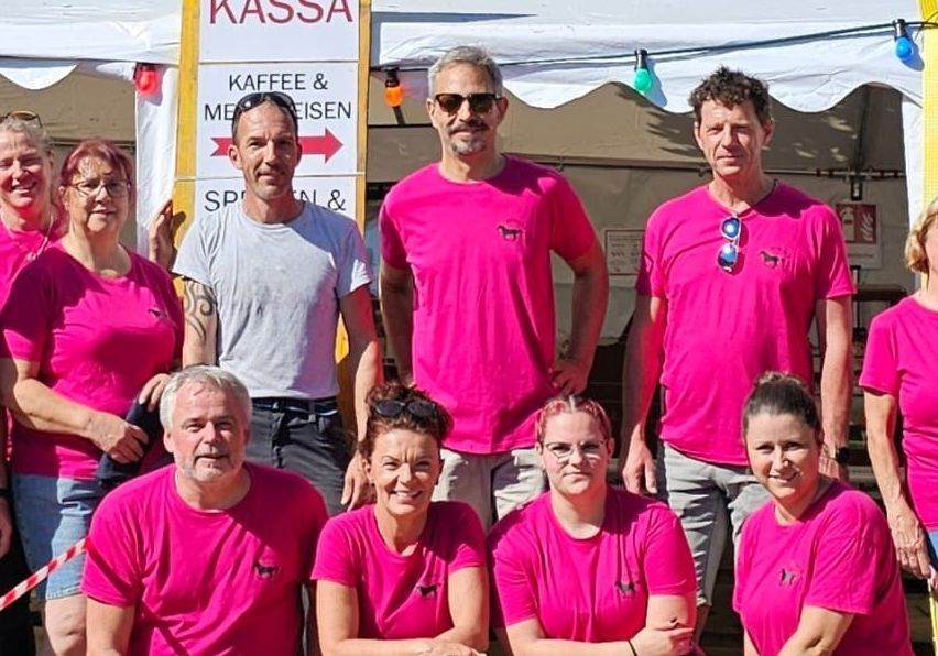 Eine Gruppe von Menschen in rosa T-Shirts posiert für ein Foto vor einem Zelt mit einem Schild, das KASSA KAFFEE & MEISIEN liest.