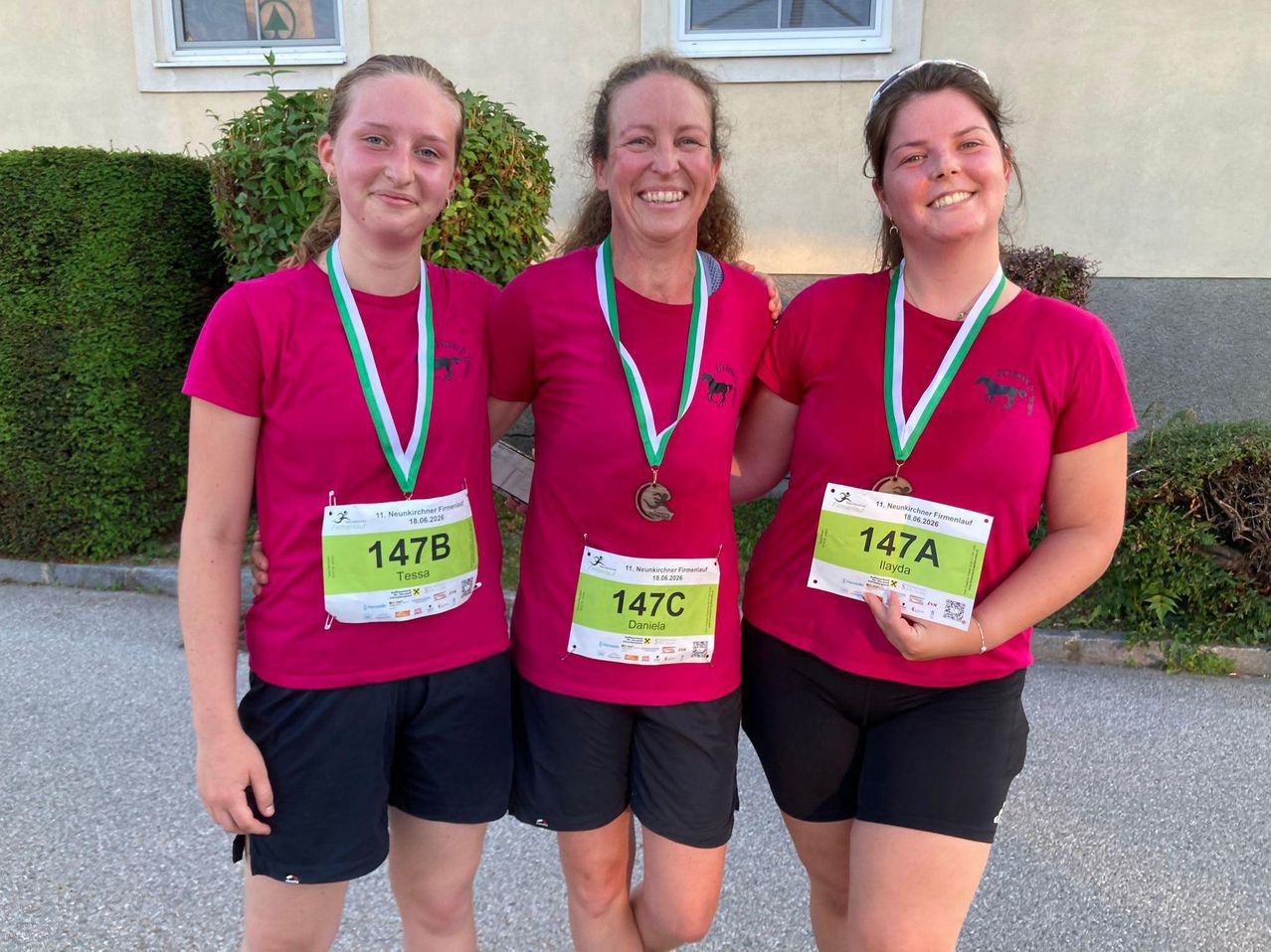 Drei Frauen in roten T-Shirts mit Pferdelogos posieren für ein Foto, mit Medaillen um den Hals. Sie lächeln, stehen in einer Reihe mit den Nummern 147B, 147C und 147A.