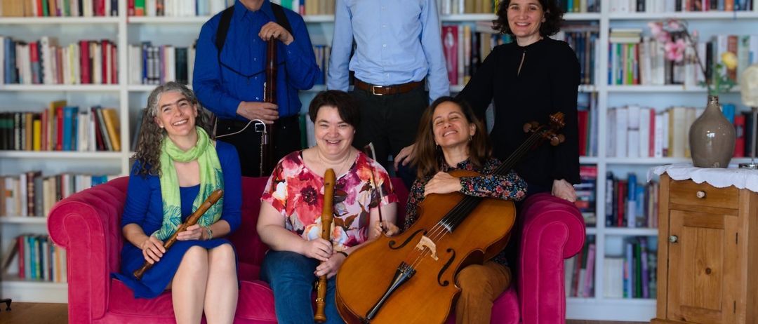 Eine Gruppe von Musikern, die verschiedene Instrumente halten, sitzen auf einem rosa Sofa vor einem Bücherregal voller Bücher.