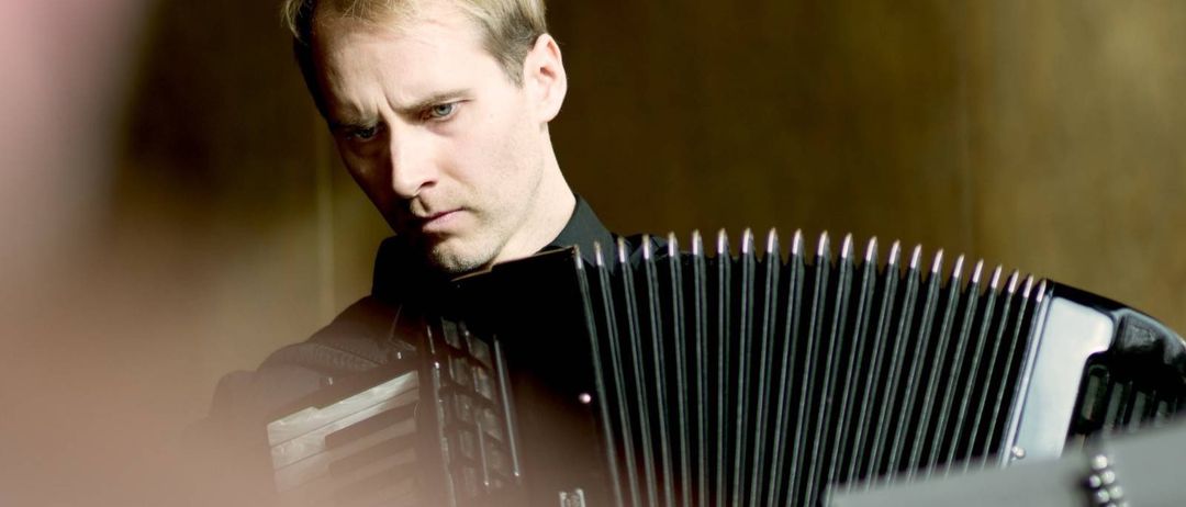 Ein Mann mit kurzen blonden Haaren spielt ein schwarzes Akkordeon und konzentriert sich intensiv auf seine Darbietung.