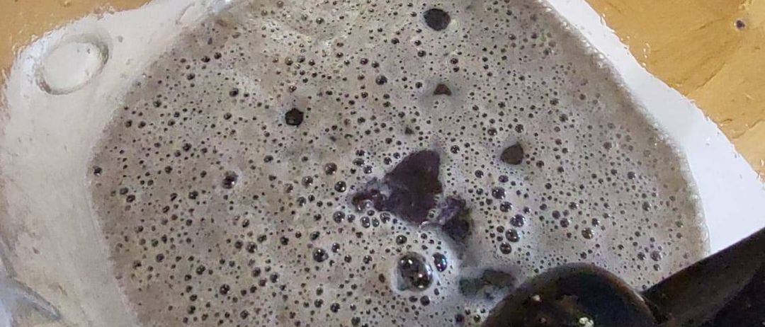 Ein Mixerbehälter mit einer Mischung aus Flüssigkeit und kleinen Blasen, auf einer Holzoberfläche platziert, wobei ein Fuß oben sichtbar ist.