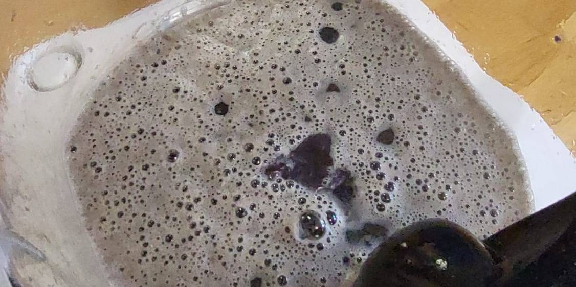 Ein Mixerbehälter mit einer Mischung aus Flüssigkeit und kleinen Blasen, auf einer Holzoberfläche platziert, wobei ein Fuß oben sichtbar ist.