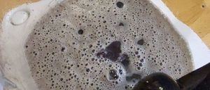 Ein Mixerbehälter mit einer Mischung aus Flüssigkeit und kleinen Blasen, auf einer Holzoberfläche platziert, wobei ein Fuß oben sichtbar ist.