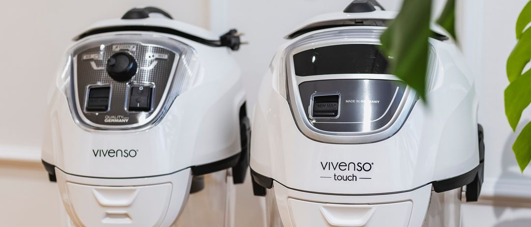 Zwei weiße Vivenso Touch Luftreiniger stehen auf einem Holztisch. Sie haben transparente Behälter und schwarze Bedienfelde. Ein grünes Blatt ist dahinter.