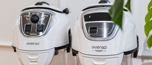 Zwei weiße Vivenso Touch Luftreiniger stehen auf einem Holztisch. Sie haben transparente Behälter und schwarze Bedienfelde. Ein grünes Blatt ist dahinter.
