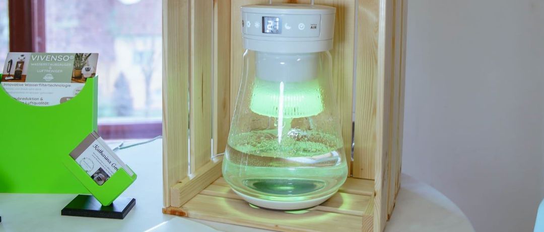 Eine Holzkiste mit einem grün beleuchteten Wasserfiltergerät steht auf einem Tisch, begleitet von drei Flaschen No-Agua. Papiere und Stifte liegen ebenfalls auf dem Tisch.