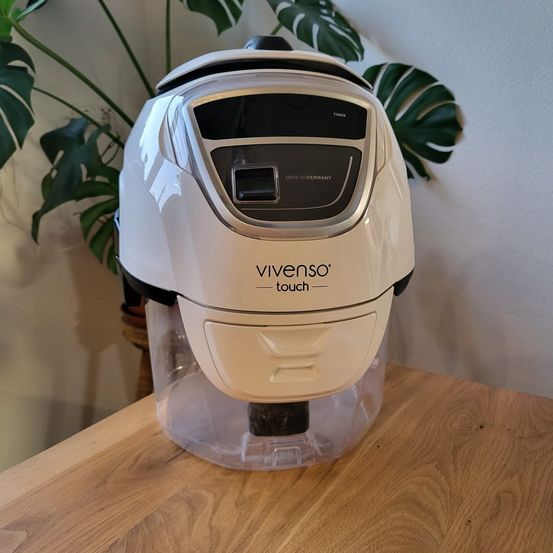 Ein weißer Vivenso Touch-Gerät, wahrscheinlich ein Luftreiniger, steht auf einem Holztisch mit einer Pflanze im Hintergrund.