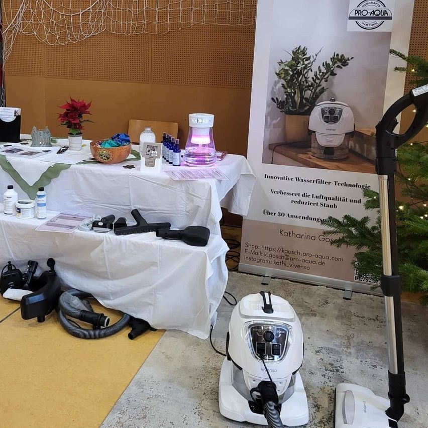 Ein Tisch mit einer weißen Tischdecke zeigt verschiedene Reinigungsprodukte und eine Pflanze. Ein Plakat mit einer Pflanze und Text steht neben dem Tisch. Ein weißer Staubsauger ist im Vordergrund.