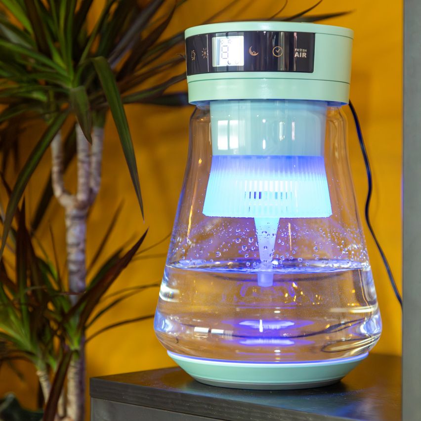 Ein zylindrischer Luftreiniger steht auf einem Tisch. Er hat einen Wassertank mit einer blauen Lampe im Inneren und einen kleinen Bildschirm oben.