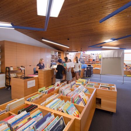 Eine Bibliothek mit Holzdecke, Regalen voller Bücher und Kindern, die Bücher halten. Eine erwachsene Frau sitzt an einem Tresen.