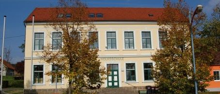 Ein beiges Gebäude mit rotem Dach und weißer Einfassung. Es hat viele Fenster und eine grüne Tür. Bäume und Gras umgeben das Gebäude.