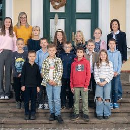 Eine Gruppe von Kindern und zwei Frauen stehen auf Stufen vor einem Gebäude mit grünen Türen. Sie posieren für ein Foto.