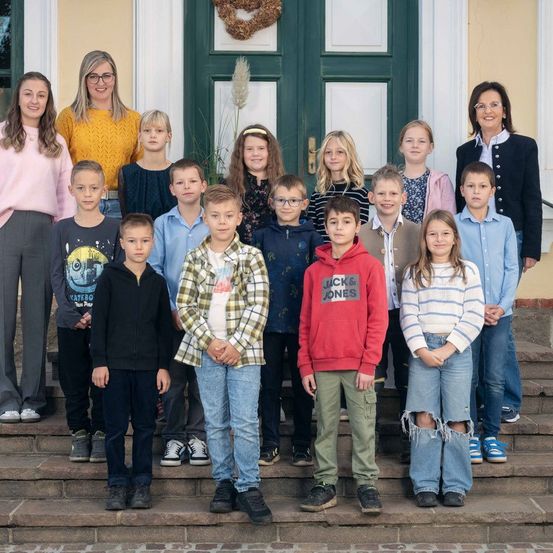 Eine Gruppe von Kindern und zwei Frauen stehen auf Stufen vor einem Gebäude mit grünen Türen. Sie posieren für ein Foto.