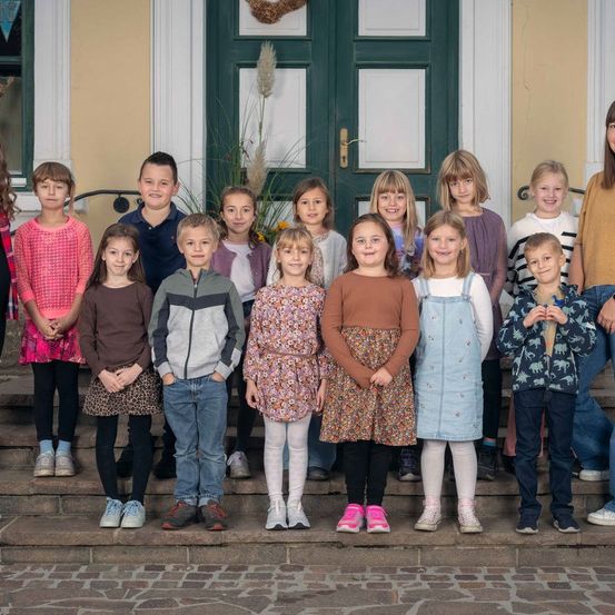 Eine Gruppe von Kindern in Winterkleidung steht vor einem Gebäude mit einem Kranz an der Tür. Sie lächeln und posieren für ein Foto.