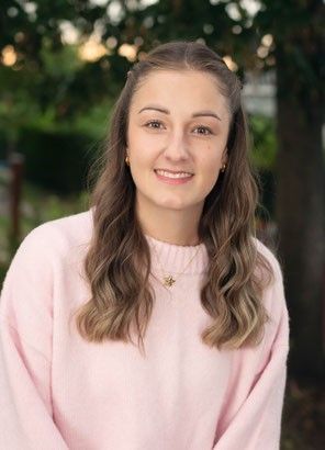Eine Frau mit langen, welligen Haaren lächelt fröhlich in einem rosa Pullover, sie steht draußen mit Bäumen im Hintergrund.