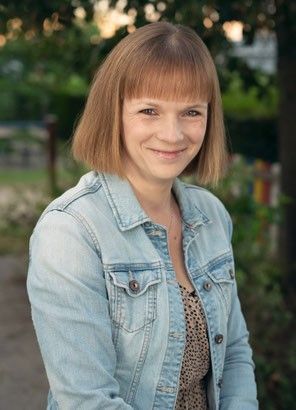 Eine Frau mit kurzen blonden Haaren steht im Freien in einer Jeansjacke und lächelt in die Kamera. Sie hat eine Halskette und ist von Bäumen und Pflanzen umgeben.