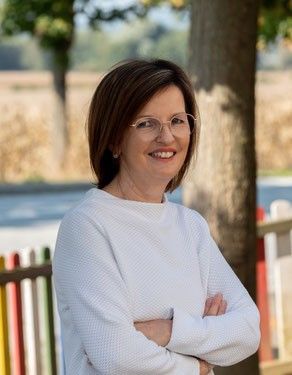 Eine Frau mit kurzen Haaren, die eine Brille und ein weißes Sweatshirt trägt, steht lächelnd mit verschränkten Armen vor einem bunten Zaun. Dahinter steht ein Baum und ein Feld.