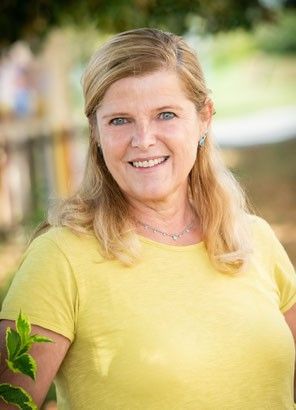 Eine Frau mit blonden Haaren steht in einem Außenbereich. Sie trägt ein gelbes Shirt und lächelt. Sie steht neben einer grünen Pflanze. Der Bereich ist von einem Holzzaun umgeben.