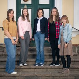 Fünf Frauen stehen auf einer Treppe vor einer grünen Tür und lächeln in die Kamera. Jede trägt verschiedene Kleidungsstücke und Schuhe und präsentiert lässige Outfits.