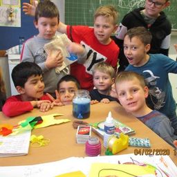 Eine Gruppe von Kindern und eine erwachsene Frau sind in einem Klassenzimmer und lächeln. Sie machen Kunst und der Tisch ist mit verschiedenen Bastelmaterialien bestückt.