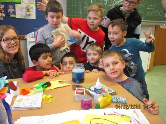 Eine Gruppe von Kindern und eine erwachsene Frau sind in einem Klassenzimmer und lächeln. Sie machen Kunst und der Tisch ist mit verschiedenen Bastelmaterialien bestückt.