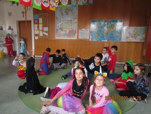 Eine Gruppe von Kindern in einem Klassenzimmer, einige in Kostümen gekleidet, sitzen auf einem grünen Kreis. Verschiedene Karten und Flaggen hängen an der Wand. Ein Datum ist in der unteren rechten Ecke.