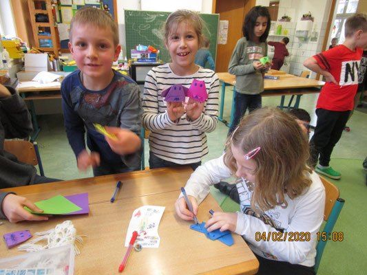 Kinder arbeiten in einem Klassenzimmer an Bastelarbeiten, eine Mädchen hält pinke Papierkreationen und eine andere malt. Sie haben Papier, Stifte und Scheren auf ihrem Tisch. Ein Junge bastelt eine Papierkreation.