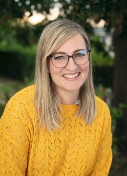 Eine Frau mit blonden Haaren, die eine Brille und einen gelben Strickpullover trägt, lächelt im Freien.
