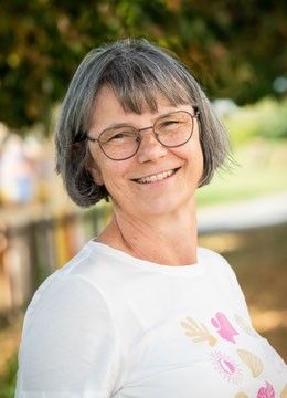 Eine ältere Frau mit kurzem grauem Haar und Brille lächelt in einem Park. Sie trägt ein weißes T-Shirt mit Blumenmuster. Bäume und ein Holzzaun sind im Hintergrund zu sehen.
