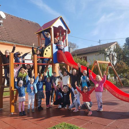 Eine Gruppe von Kindern in Winterkleidung posiert für ein Foto auf einem Spielplatz mit Rutschen und Schaukeln, vor dem Hintergrund eines Gebäudes und Bäumen.