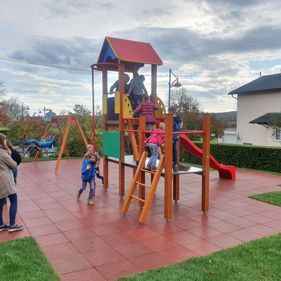 Ein Spielplatz mit Rutsche und Klettergerüst. Kinder spielen und einige Erwachsene stehen herum.