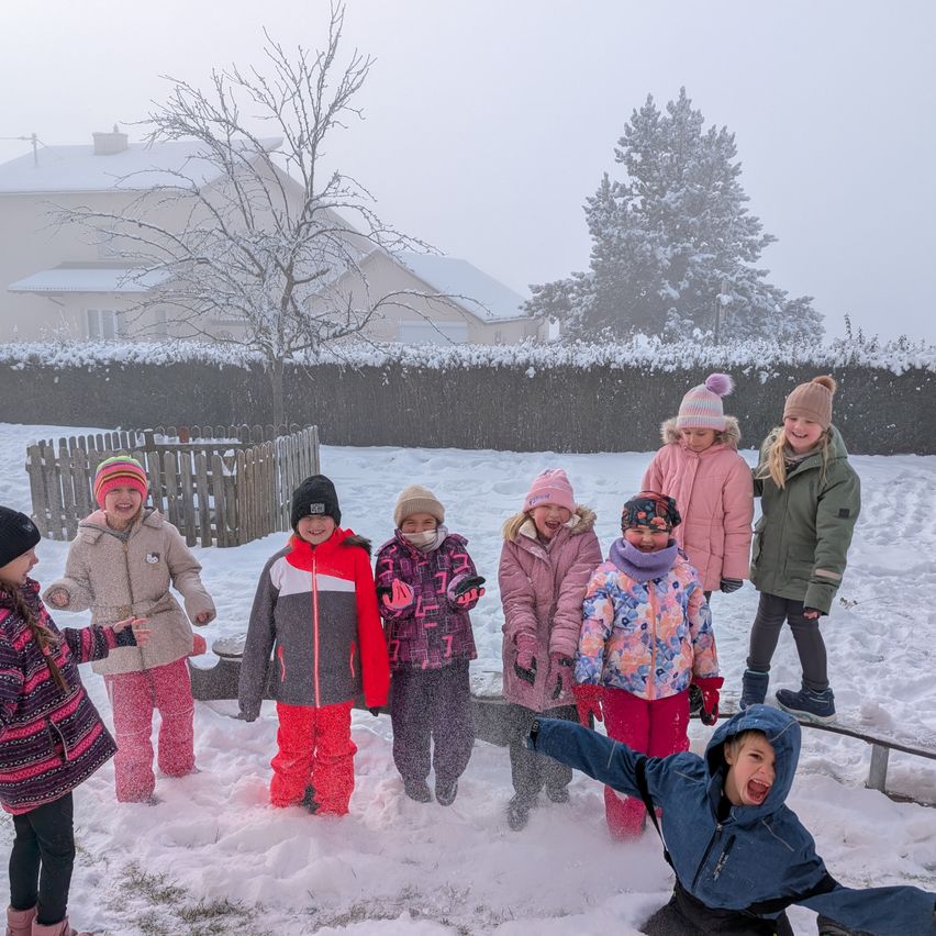 Eine Gruppe von Kindern spielt im Schnee im Hinterhof, angezogen in Winterkleidung. Im Hintergrund sieht man ein Haus und einen Zaun.