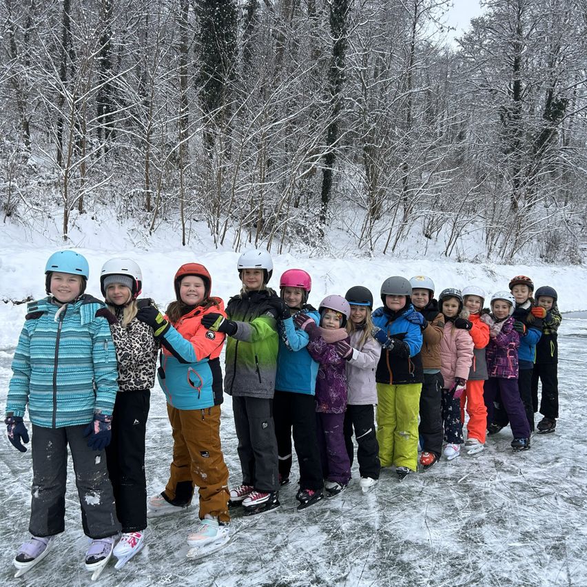 Eine Gruppe von Kindern in Winterkleidung steht auf einer Eisbahn und lächelt für ein Foto. Sie tragen Helme und verschiedene farbige Winterkleidung. Der Hintergrund zeigt schneebedeckte Bäume.