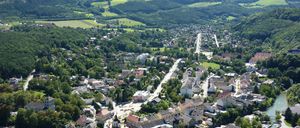 Ein Luftbild einer Stadt, eingebettet zwischen grünen Hügeln, mit Gebäuden verschiedener Größen und Stilen, umgeben von üppigen Bäumen und einem mäandernden Fluss.