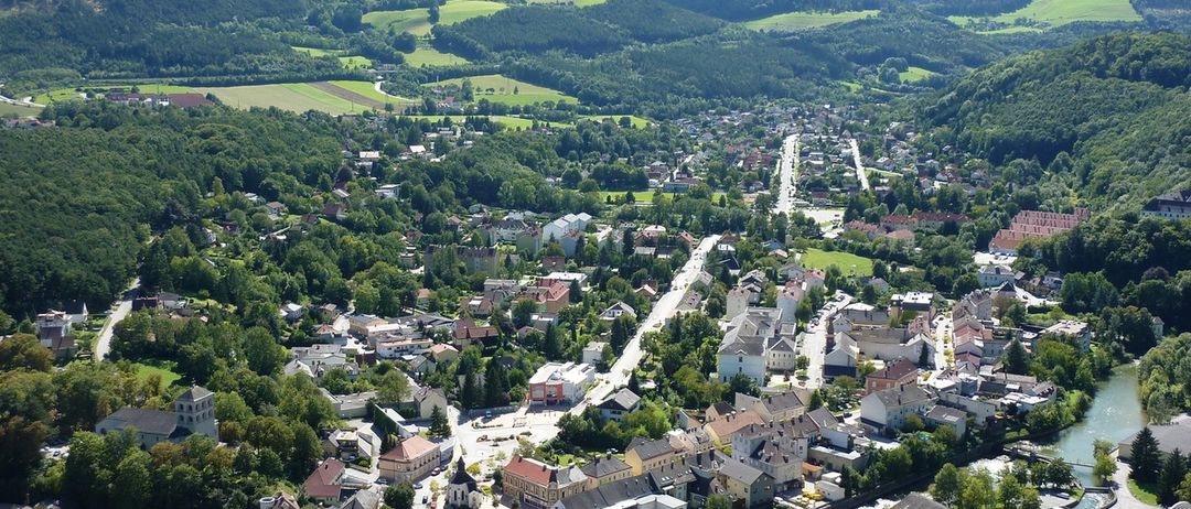 Ein Luftbild einer Stadt, eingebettet zwischen grünen Hügeln, mit Gebäuden verschiedener Größen und Stilen, umgeben von üppigen Bäumen und einem mäandernden Fluss.
