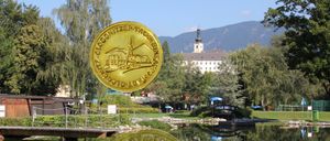 Eine goldene Münze mit Schlossdesign schwebt über einem ruhigen See. Im Hintergrund steht ein großes Gebäude mit Turm vor einer Bergkulisse.