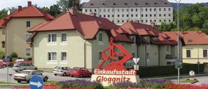 Ein großes Gebäude mit einem Turm und vielen Fenstern steht hinter einem farbenfrohen Blumenbeet. Ein Schild mit den Worten Einkaufsstadt Gloggnitz steht davor. Autos sind vor dem Gebäude geparkt.