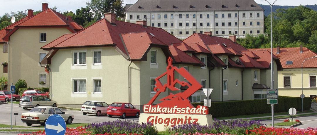 Ein großes Gebäude mit einem Turm und vielen Fenstern steht hinter einem farbenfrohen Blumenbeet. Ein Schild mit den Worten Einkaufsstadt Gloggnitz steht davor. Autos sind vor dem Gebäude geparkt.