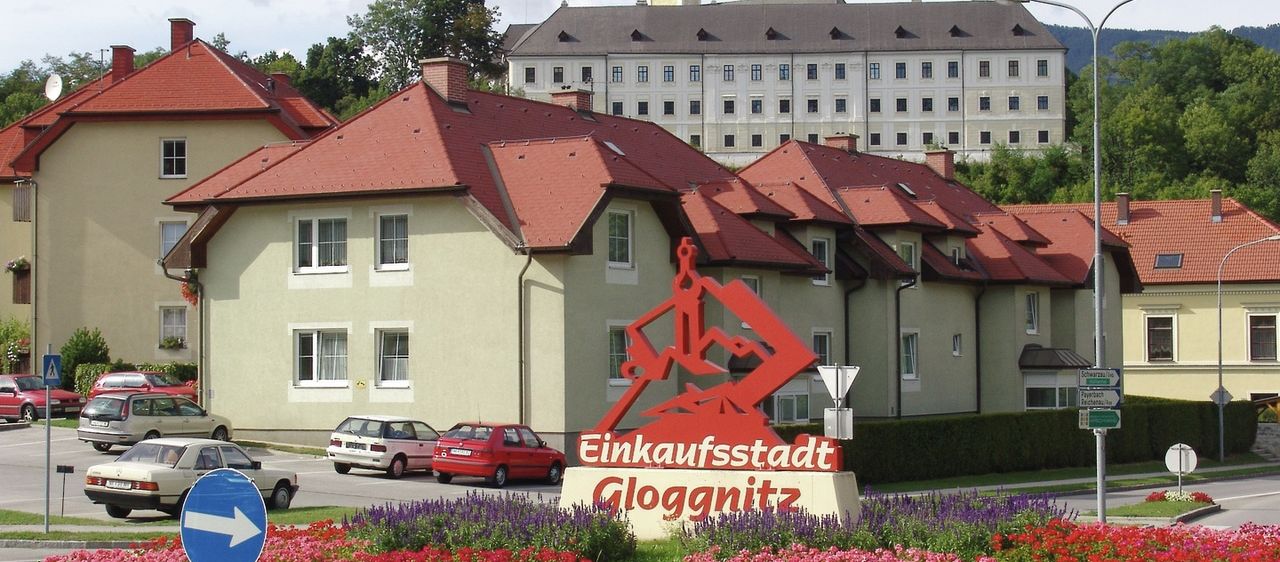 Ein großes Gebäude mit einem Turm und vielen Fenstern steht hinter einem farbenfrohen Blumenbeet. Ein Schild mit den Worten Einkaufsstadt Gloggnitz steht davor. Autos sind vor dem Gebäude geparkt.