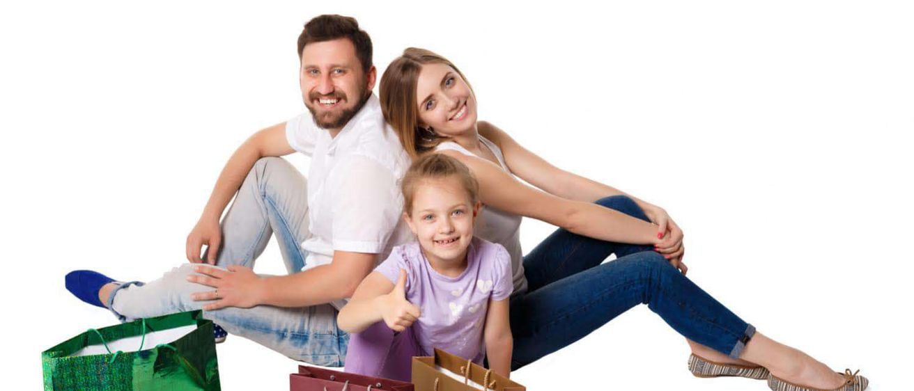 Eine lächelnde Familie aus drei Personen, ein Mann, eine Frau und ein Mädchen, umgeben von farbenfrohen Einkaufstüten, sitzt auf einem weißen Hintergrund.