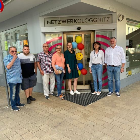 Sechs Personen stehen vor einem Geschäft namens 'Netzwerkgloggnitz'. Sie lächeln und posieren für ein Foto. Bunte Luftballons sind hinter ihnen.