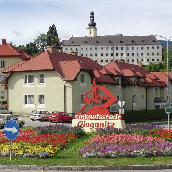Ein farbenfrohes Eingangsschild zur Einkaufsstadt Gloggnitz, das eine rote gebirgige Figur zeigt, umgeben von bunten Blumen. Dahinter ein großes Gebäude mit roten Dächern und vielen Fenstern.