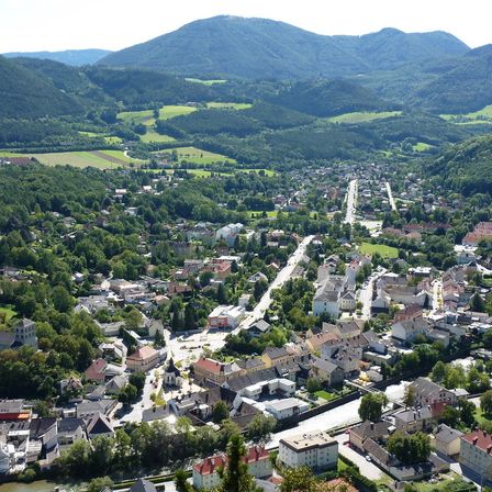 Ein Luftbild einer Stadt im Tal mit dichten Bäumen und Gebäuden, umgeben von grünen Hügeln und Bergen in der Ferne.