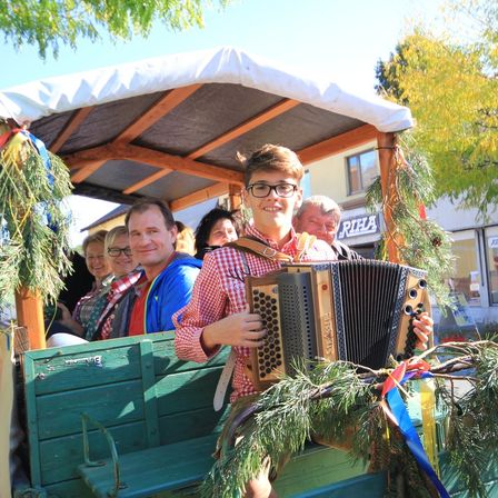 Ein Junge spielt ein Akkordeon in einem dekorierten Wagen, mit Menschen dahinter. Der Wagen hat ein Dach und steht auf der Straße. Hinter dem Wagen befindet sich ein Gebäude mit einem Schild, auf dem 'RIFA' steht.