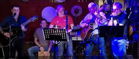 Drei Männer auf einer Bühne, einer spielt Gitarre, ein anderer Schlagzeug und der dritte ebenfalls Gitarre. Ein Mikrofon auf einem Ständer steht zwischen ihnen.