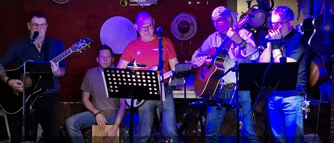 Drei Männer auf einer Bühne, einer spielt Gitarre, ein anderer Schlagzeug und der dritte ebenfalls Gitarre. Ein Mikrofon auf einem Ständer steht zwischen ihnen.