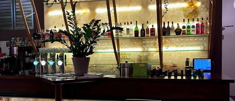 Eine Bar mit einer Holztheke, drei schwarzen Hockern, einem Monitor und einer Topfpflanze. Regale mit Flaschen und Gläsern sind mit warmen gelben Lichtern beleuchtet.