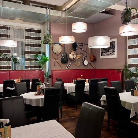 Ein leeres Restaurant mit Holzböden, Tischen, Stühlen und Lampen. Musikinstrumente hängen an der Wand. Es gibt auch Pflanzen und einen Bilderrahmen.