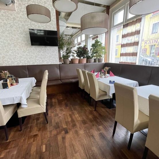 Ein leerer Essbereich mit Holzböden, Tischen und Stühlen. Topfpflanzen und ein Fernseher an der Wand.