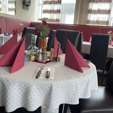 Ein Restauranttisch mit roten Servietten, Besteck und einer Blumenvase. Es gibt Stühle um den Tisch herum. Im Hintergrund befinden sich weitere Tische und ein Fenster mit Vorhängen.